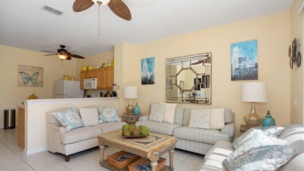 A living room with two sofas, a coffee table, a ceiling fan, and a kitchen area in the background.