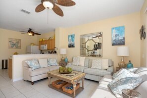 Un salon avec deux canapés, une table basse, un ventilateur de plafond et, au fond, un coin cuisine.