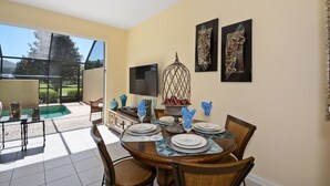 A dining area with a table set for four, a television on the wall, and a view of a swimming pool and greenery outside.