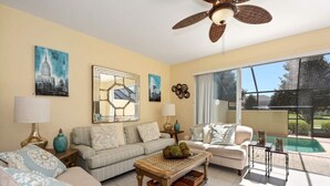 A living room with a sofa, armchairs, a coffee table, and a ceiling fan. There are paintings on the wall, a window with a view of a pool, and a door leading outside.