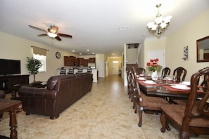 A spacious living room with a brown leather sofa, a dining area with wooden chairs and a table set for four, and a kitchen in the background. - Champions Gate IHR 3017 (Davenport)