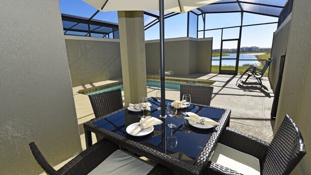 An outdoor patio with a glass table set for dining, surrounded by wicker chairs, and overlooking a swimming pool and landscape.