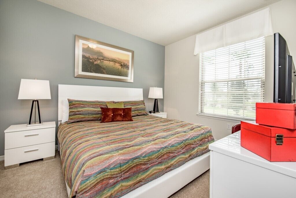 A bedroom with a bed, two bedside tables, a nightstand, a window with blinds, a painting on the wall, and a red storage box.