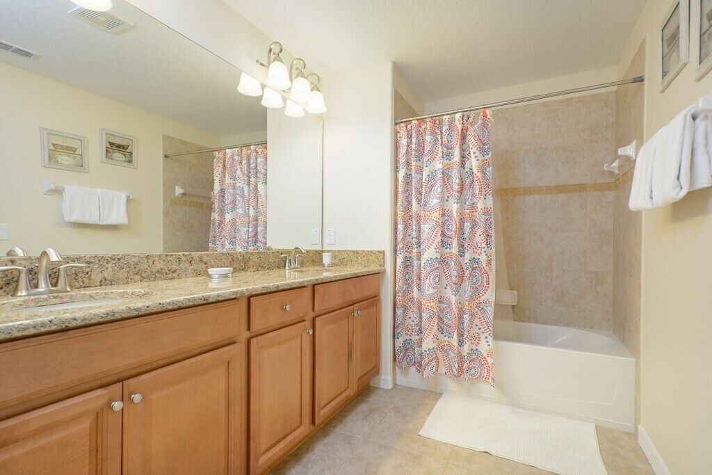 A bathroom with a large mirror, vanity with a granite countertop, wooden cabinets, a shower with a patterned curtain, and a white bathtub.