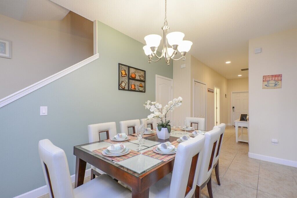 A dining area with a rectangular table set for six, surrounded by white chairs. A chandelier hangs from the ceiling, and a decorative wall piece is visible.