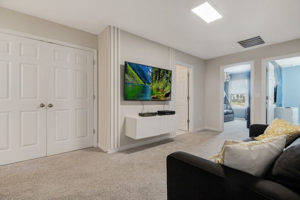 A modern living room with a flat-screen TV mounted on a white wall, a black sofa with cushions, and a carpeted floor.