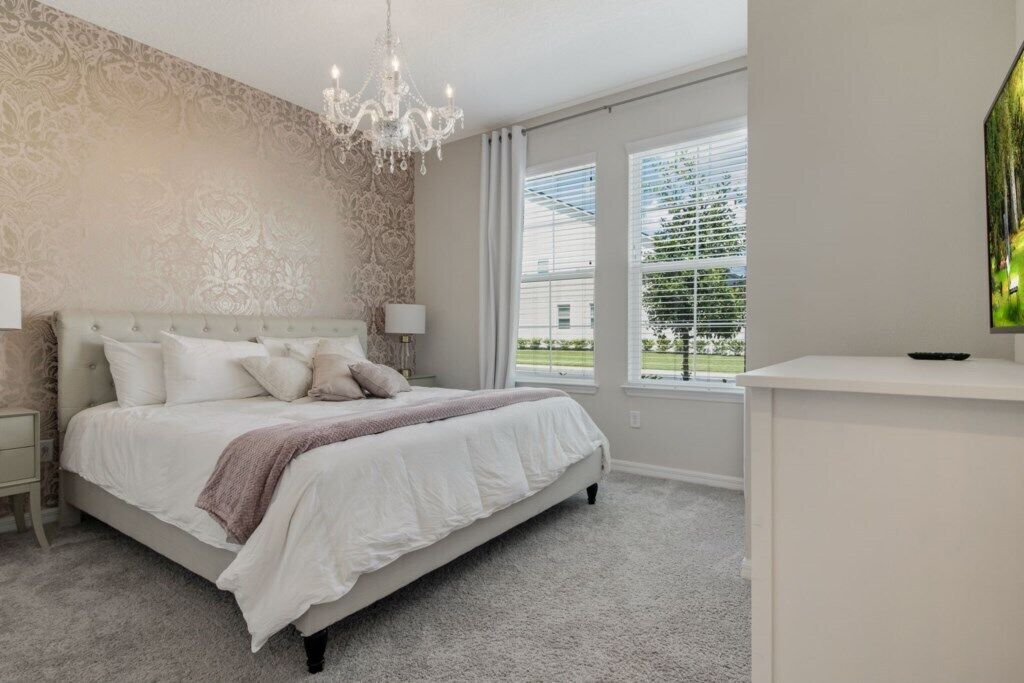 A bedroom with a large bed, a chandelier, a nightstand, a dresser, and a window with blinds.