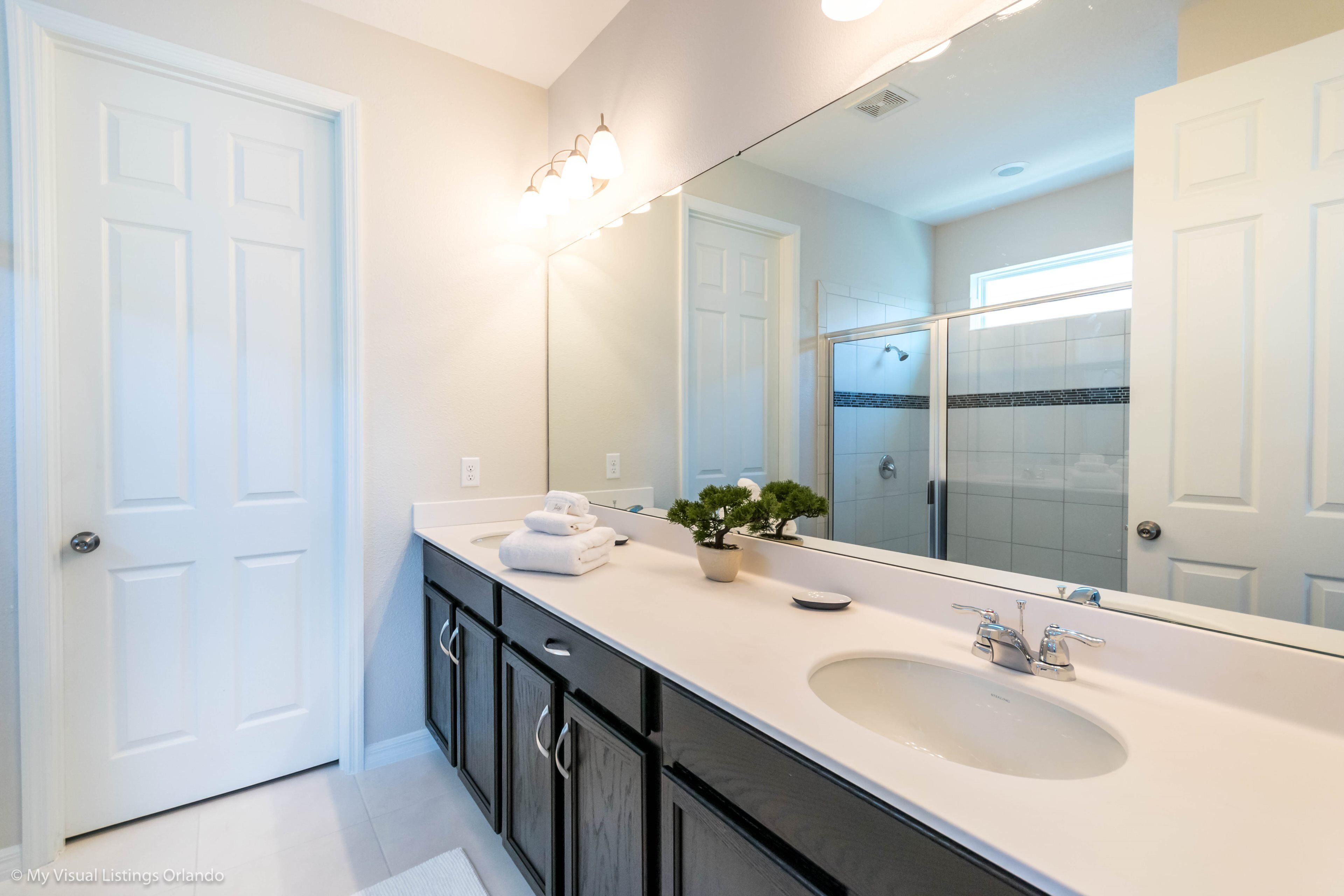 Un bagno moderno con un grande specchio, un mobile lavabo bianco, pensili scuri e una pianta in vaso.