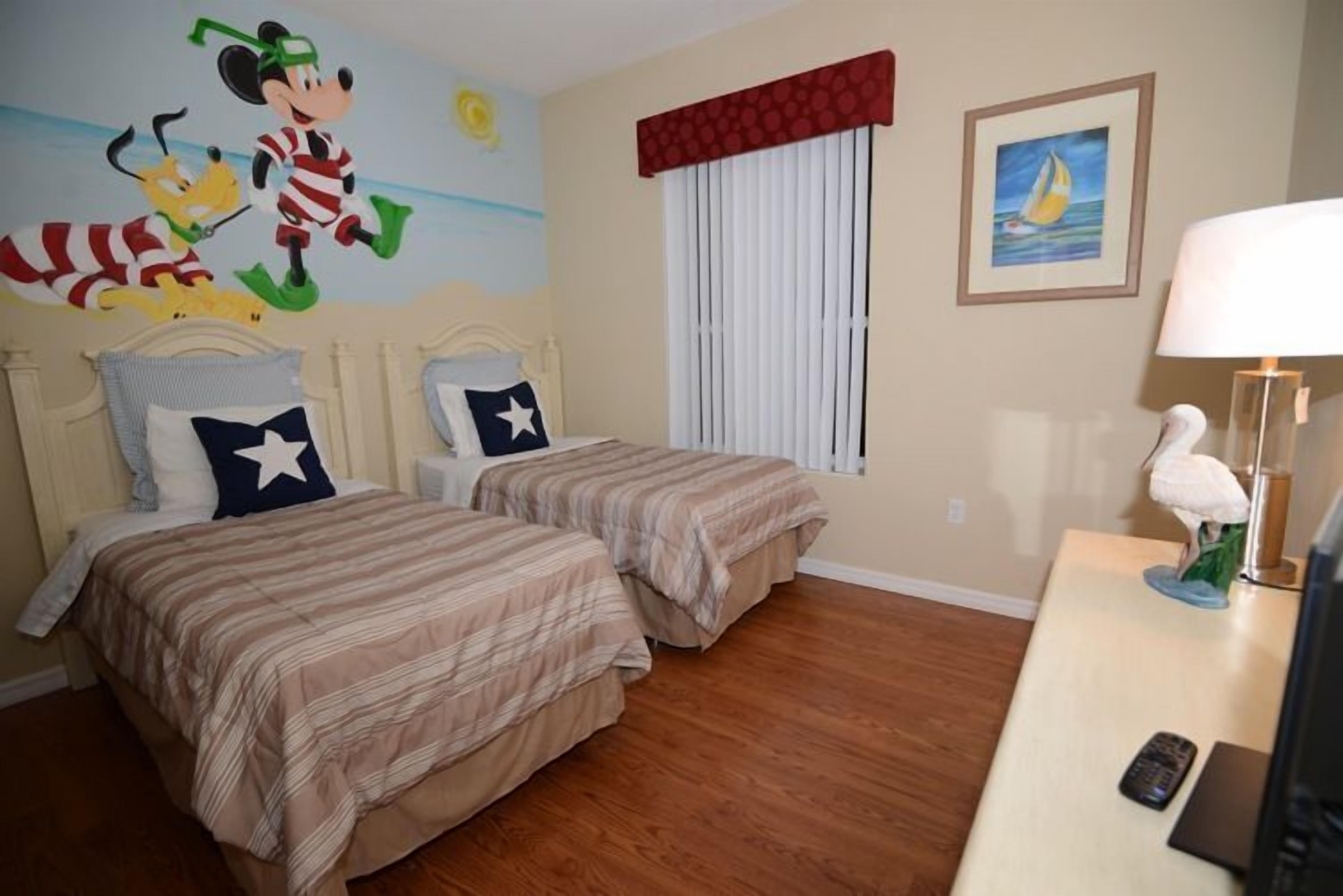 A hotel room with two beds, a mural of cartoon characters, a sailboat painting, and a desk with a lamp and remote control.