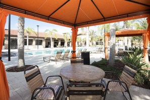 A covered outdoor seating area with a pool in the background.