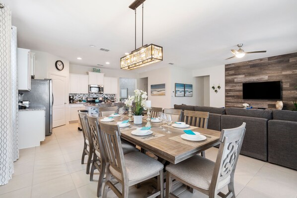 A modern kitchen with white cabinets, a built-in oven, and a microwave. There is a dining area with a wooden table set for four, surrounded by chairs. A large window provides natural light, and a flat-screen TV is mounted on the wall.