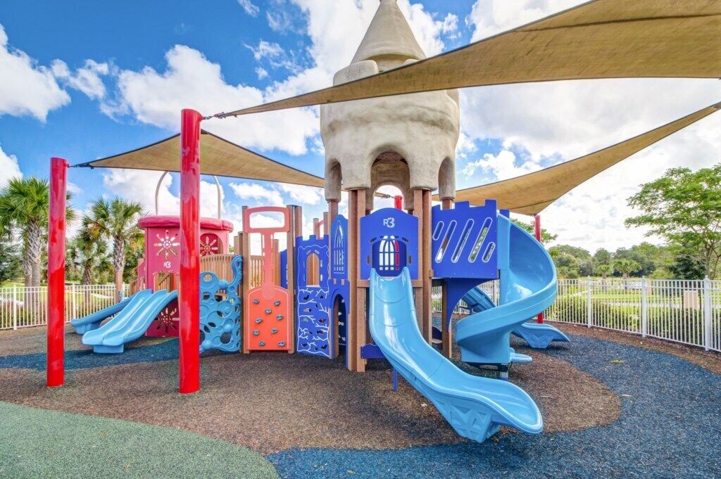 A playground with multiple slides and climbing structures, surrounded by a fence and palm trees.