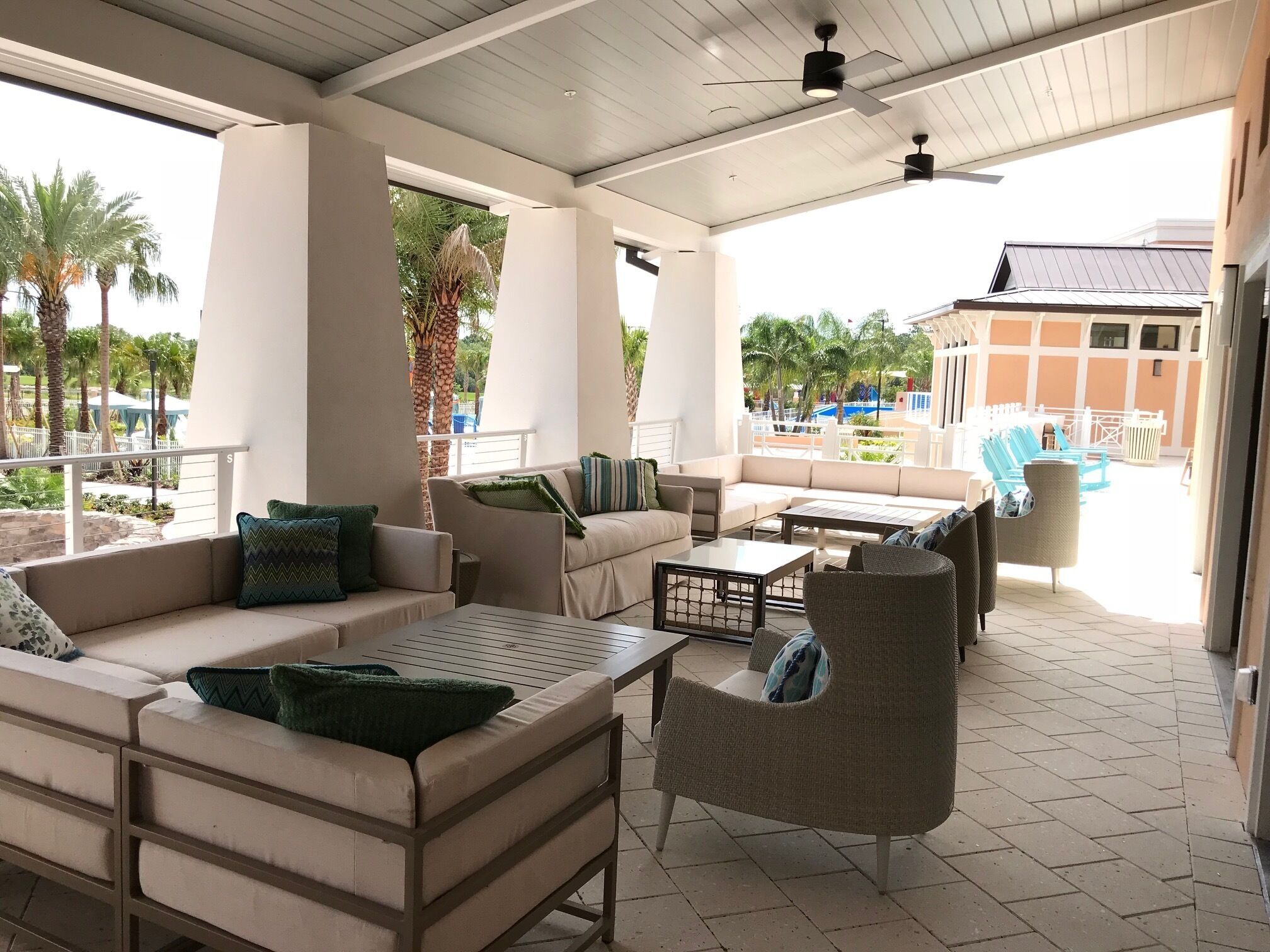 A covered outdoor seating area with sofas and chairs, a ceiling fan, and a view of a pool and palm trees.