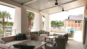 A covered outdoor seating area with sofas and chairs, a ceiling fan, and a view of a pool and palm trees.