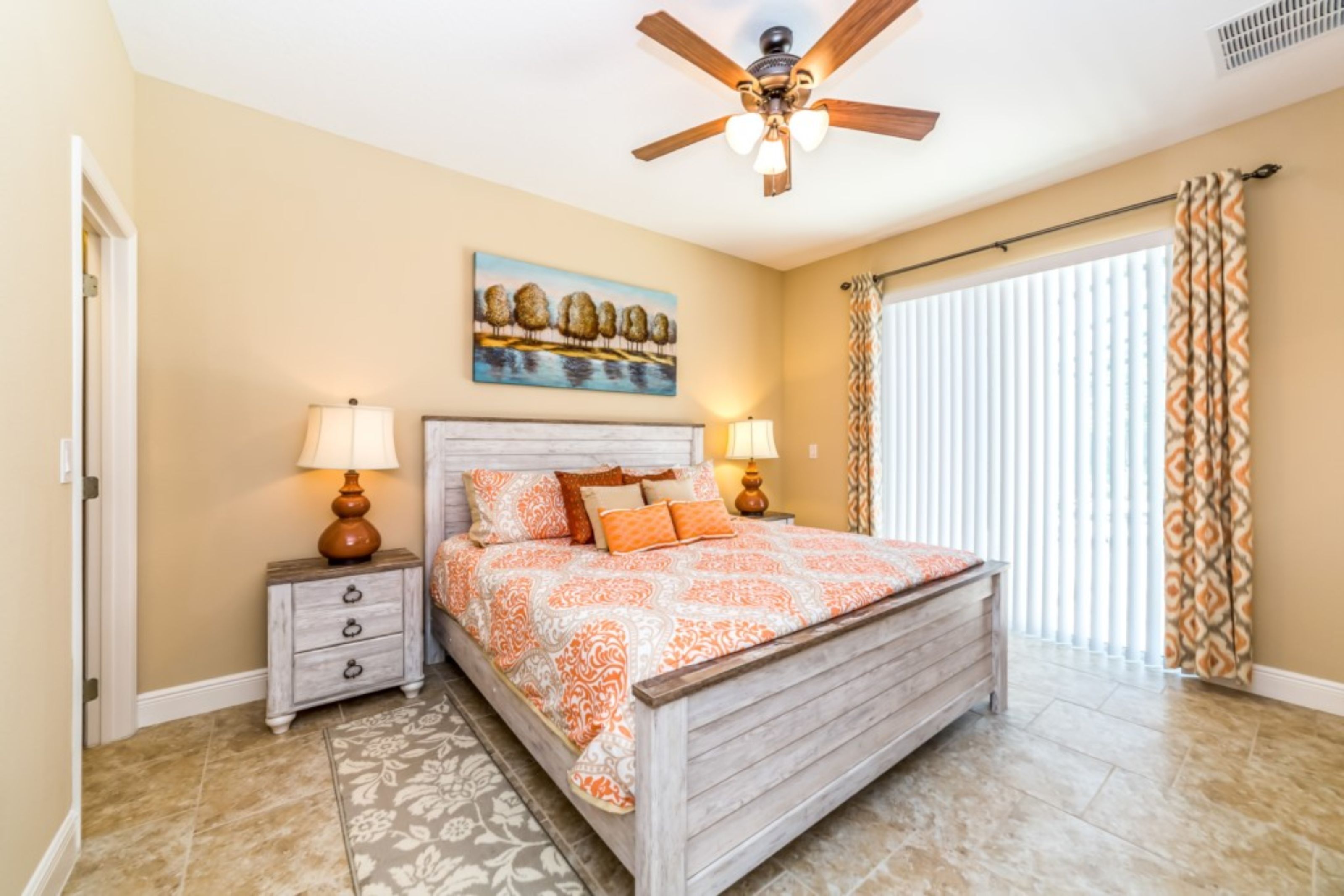 A bedroom with a wooden bed, two bedside tables with lamps, a ceiling fan, a window with blinds, and a painting on the wall.