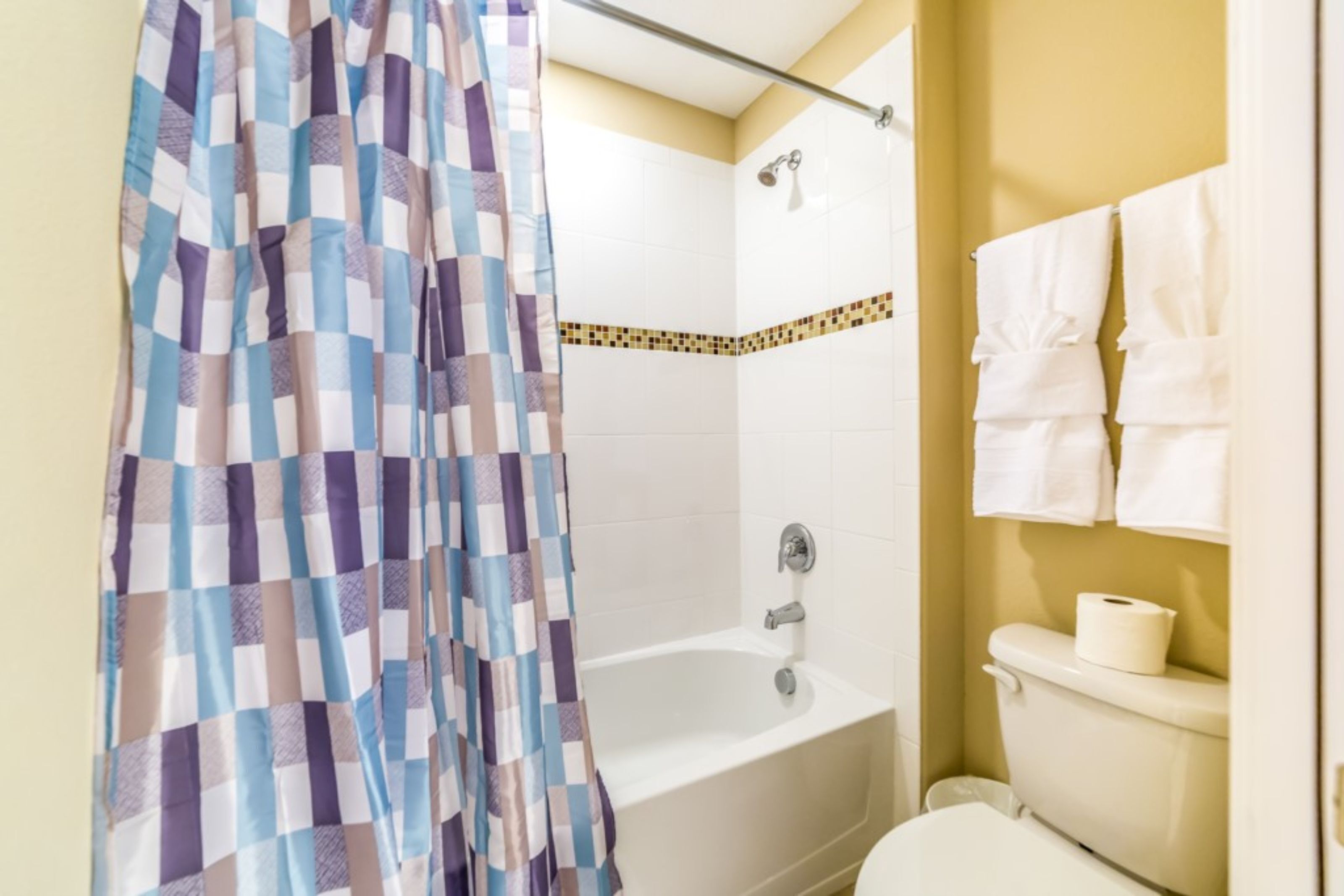 Een badkamer met een douchegordijn met ruitpatroon, een badkuip, een toilet en een handdoekrek met handdoeken.