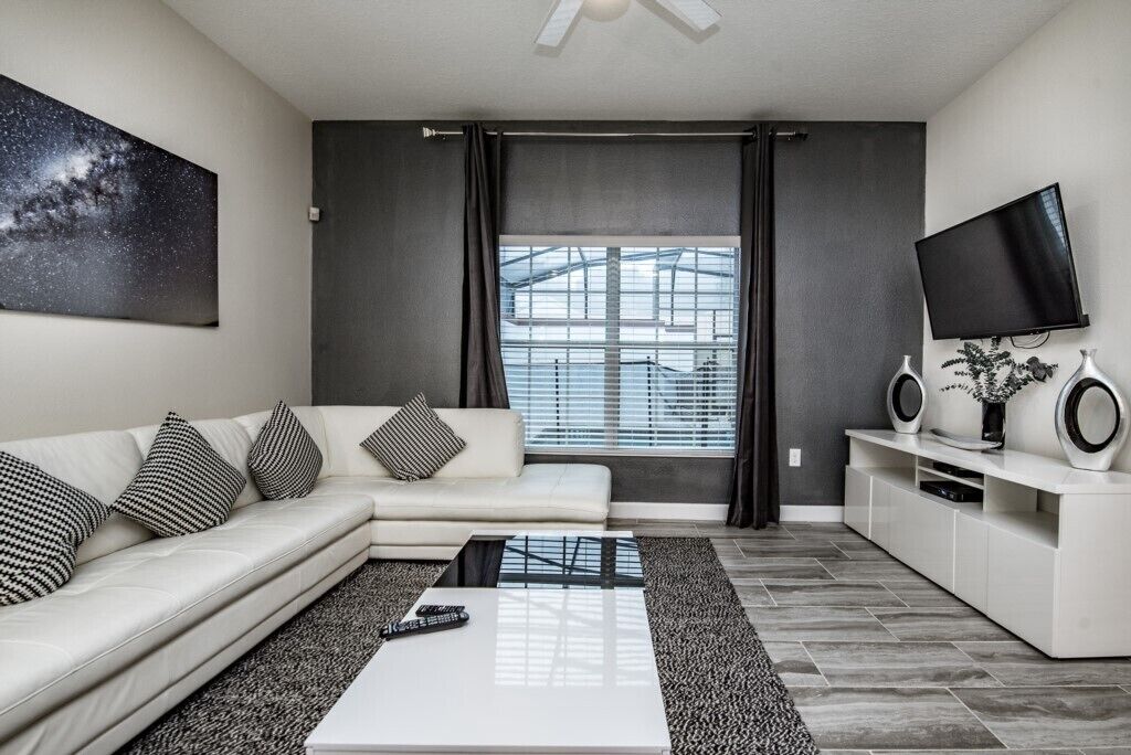 A modern living room with a sectional sofa, a coffee table, a TV, and a large window with blinds.
