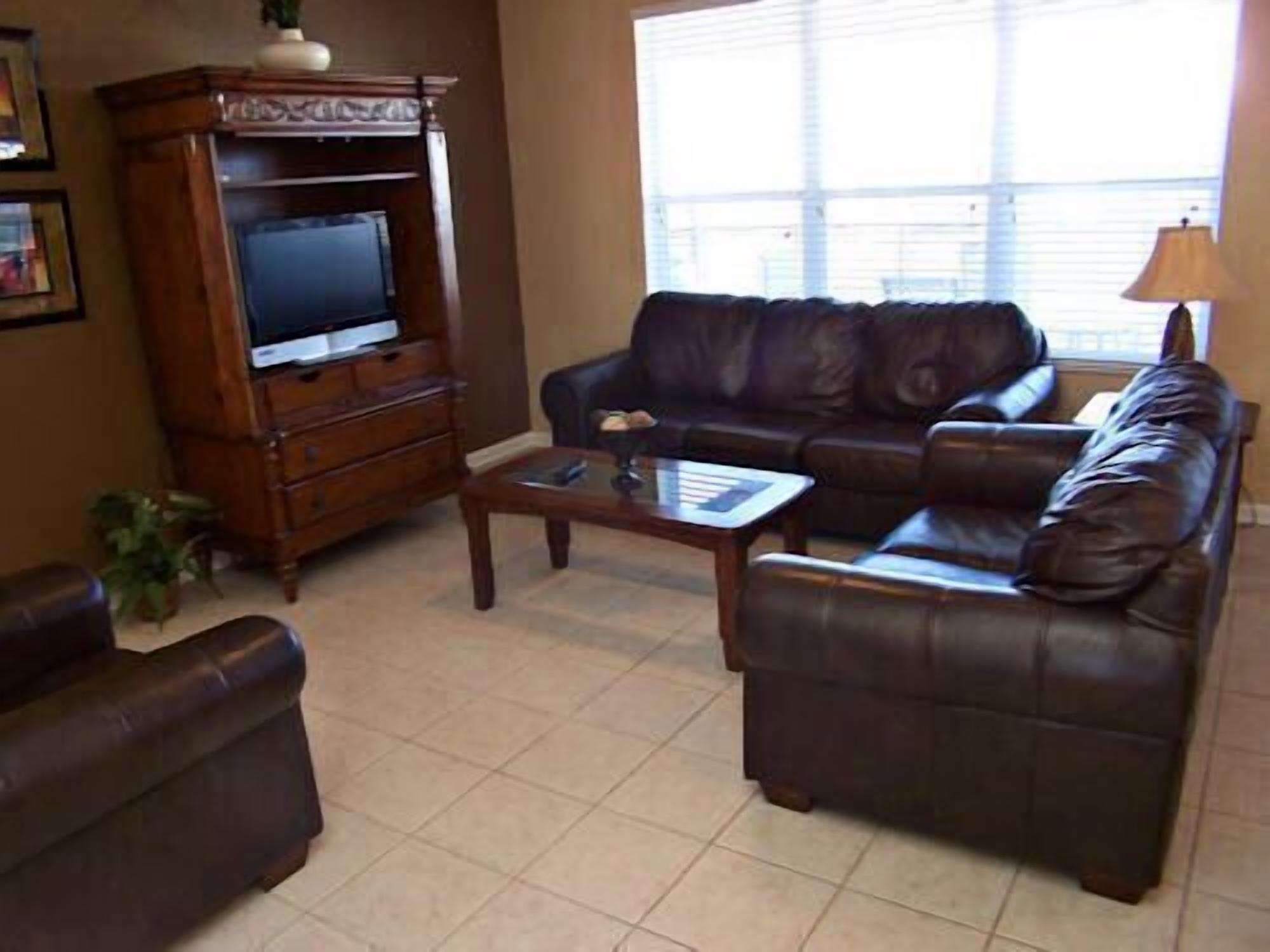 A living room with a brown leather sofa, a wooden entertainment center with a TV, and a window with blinds.