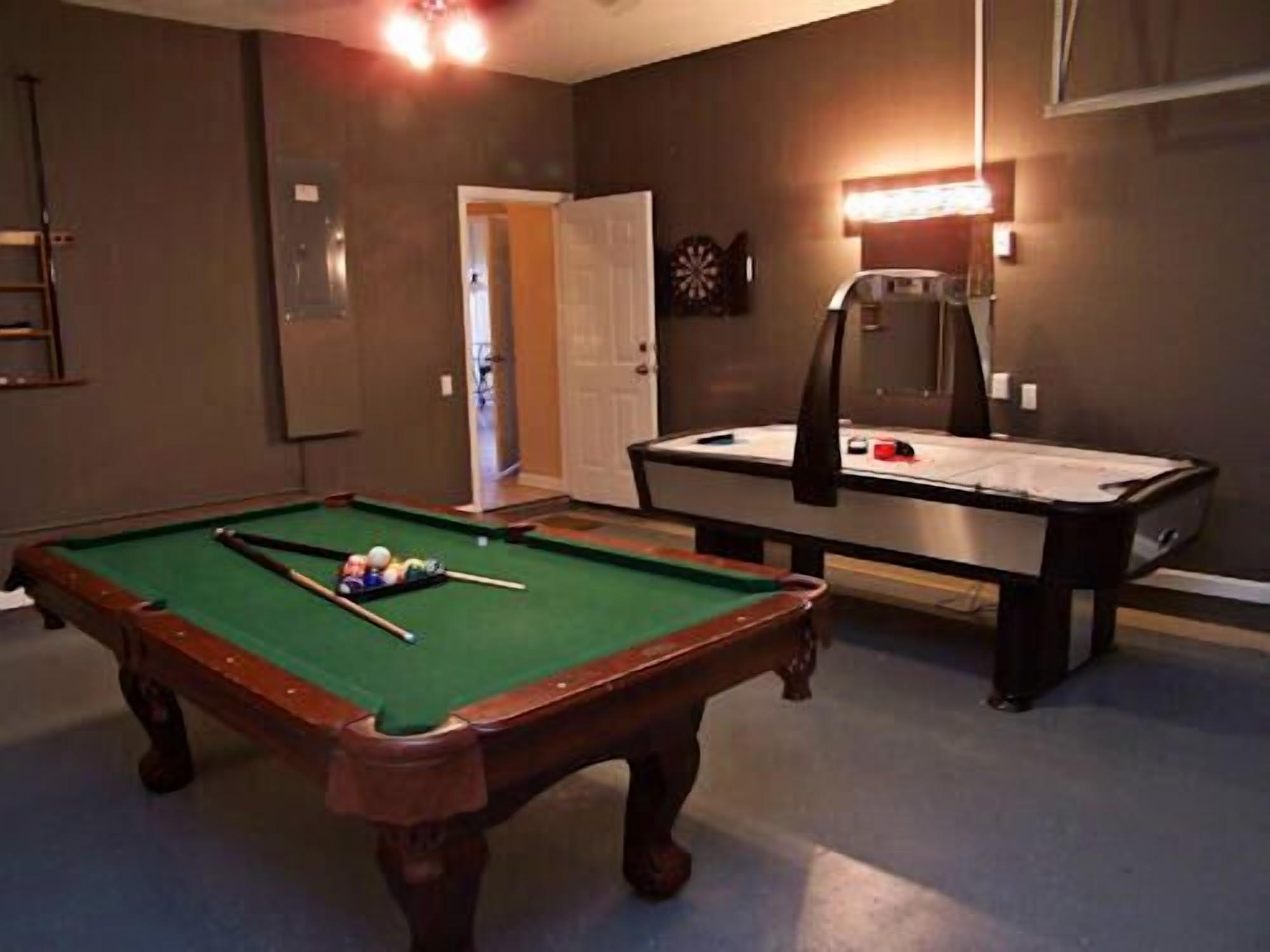 A pool table with a billiard cue and balls, a billiard rack, and a dartboard in a room with a door and a light fixture.