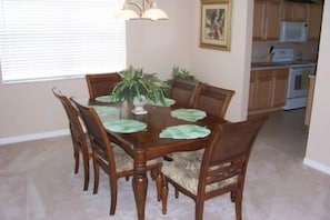 A dining room with a wooden table and chairs, a potted plant, and a kitchen in the background.