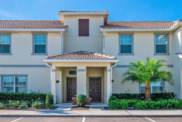 A two-story building with a tiled roof, a central entrance with two doors, and a palm tree in the foreground. - Beautiful 4 Bdrm, 3 Bath Townhome (Kissimmee)