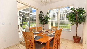 Uma sala de jantar com mesa e cadeiras de madeira, uma planta em vaso e vista para uma estufa através de grandes janelas.