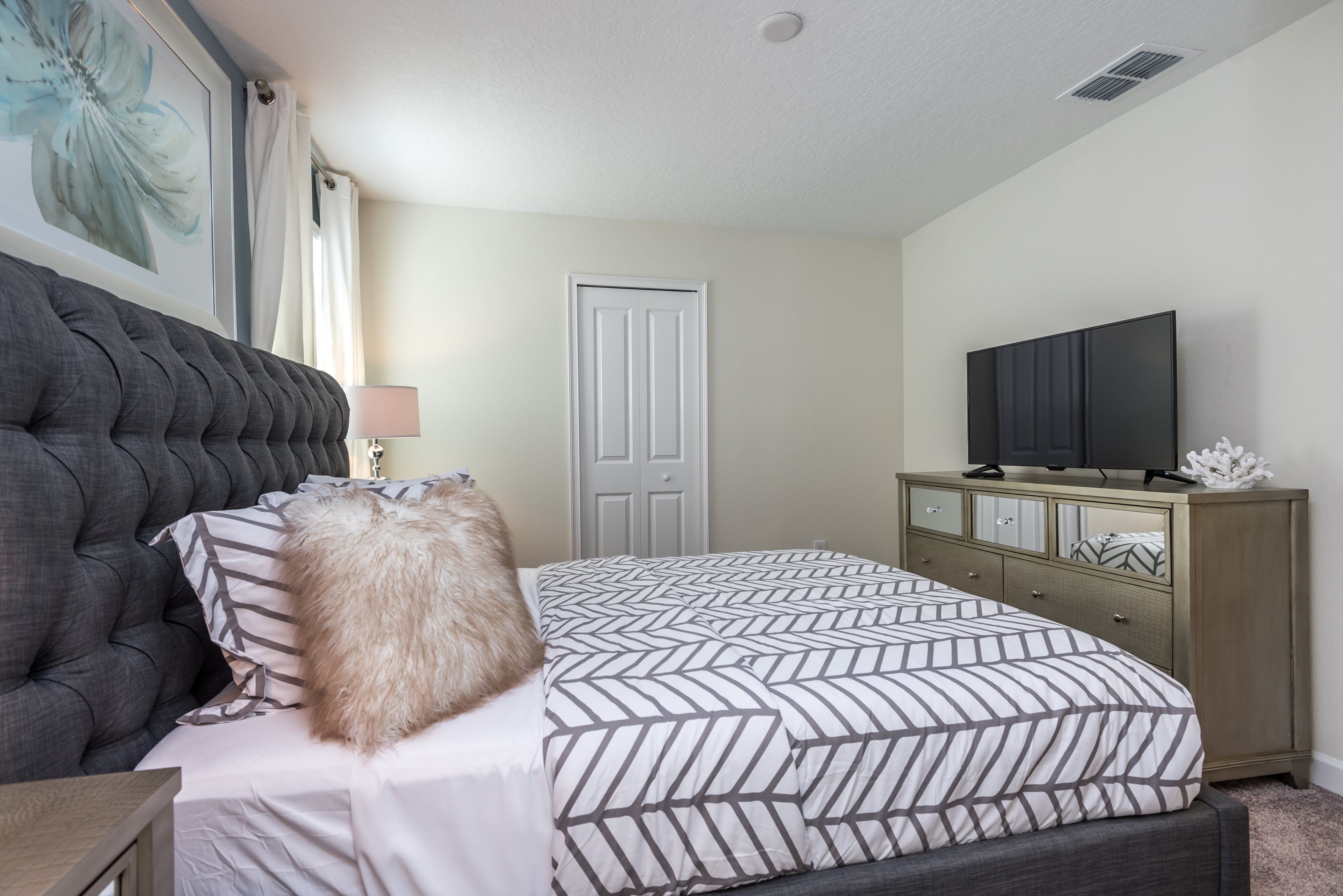 A bedroom with a tufted headboard, a bed with a patterned bedspread, a television on a cabinet, and a door in the background.