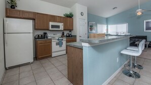 A modern kitchen with wooden cabinets, a white refrigerator, a microwave, and a stove. There is a bar area with two white bar stools and a ceiling fan. - West Haven IHR 4079 (Davenport)
