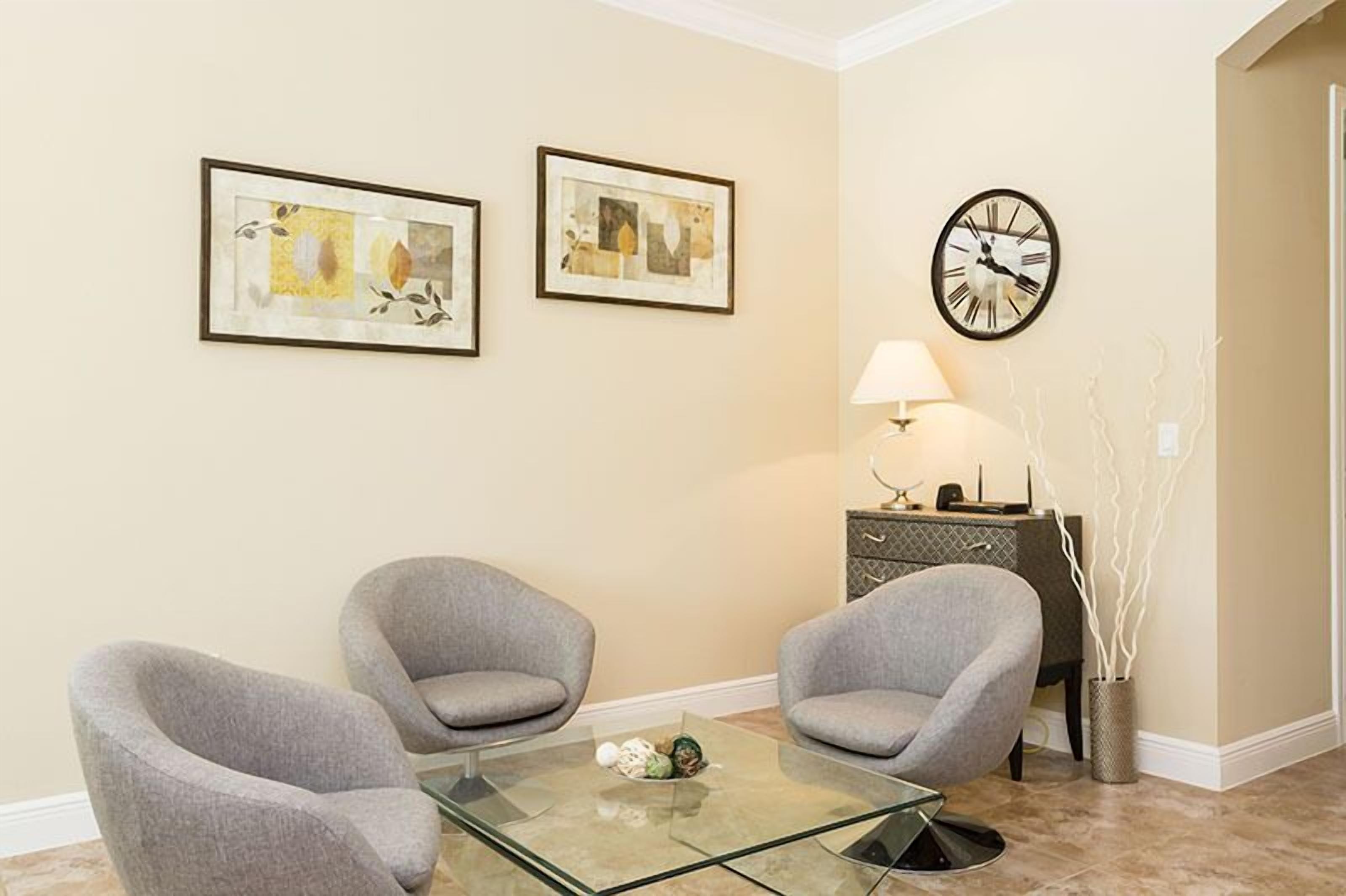 Una sala de estar moderna con dos sillones grises, una mesa de centro de vidrio, un reloj y dos cuadros abstractos enmarcados en la pared.