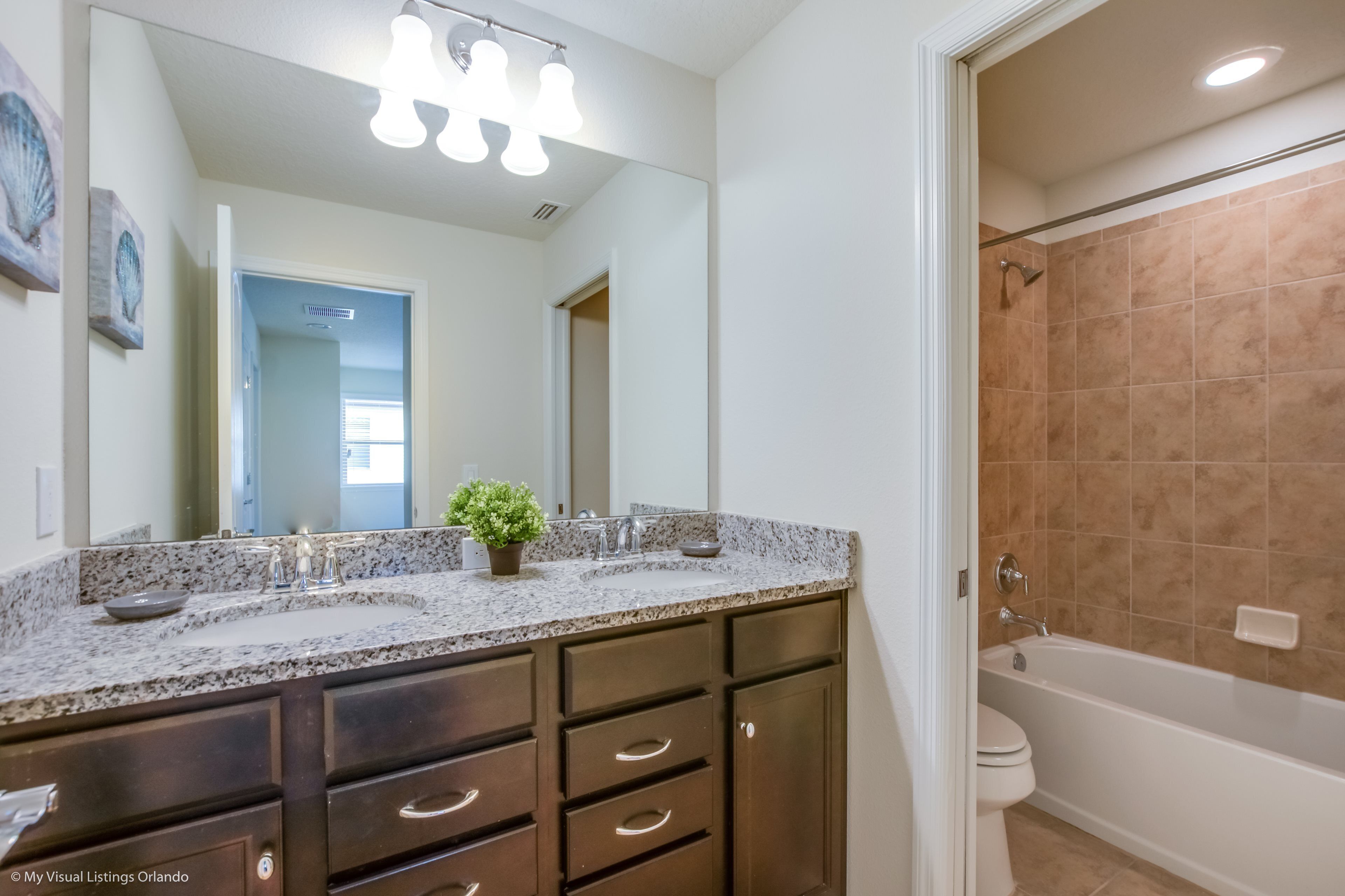 A bathroom with a double sink vanity, a large mirror, a shower with beige tiles, and a toilet.