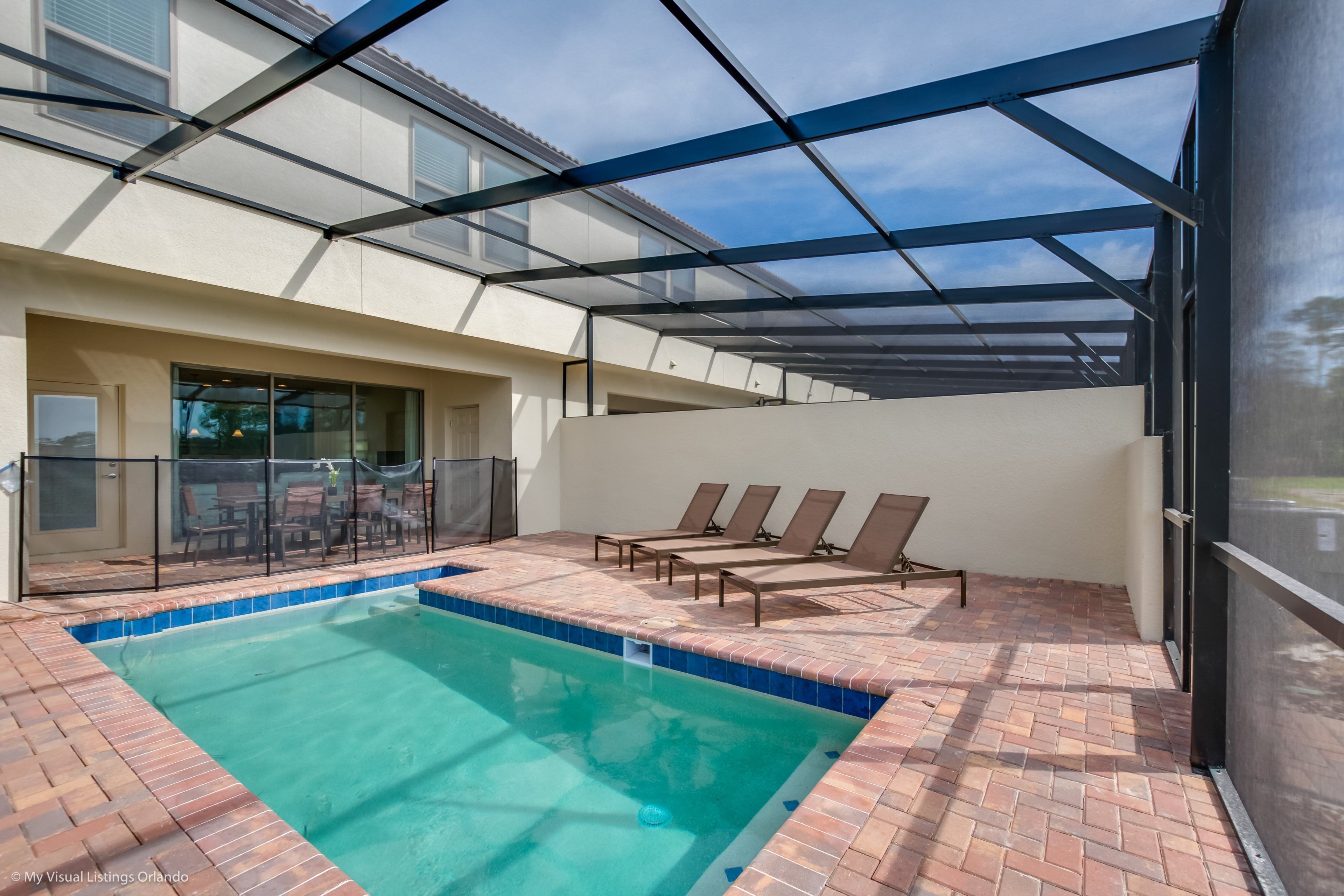 A swimming pool with a glass enclosure, sun loungers, and a patio area with a brick floor.