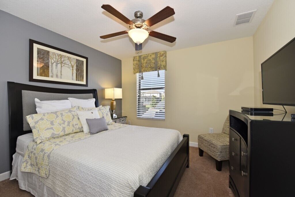 A bedroom with a bed, a ceiling fan, a window with blinds, a TV, and a dresser.