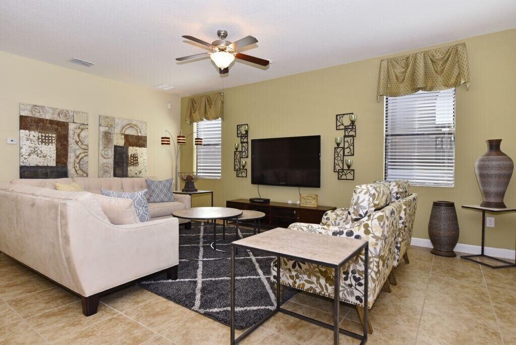 A living room with a sofa, armchairs, a coffee table, a flat-screen TV, decorative wall art, and a ceiling fan.