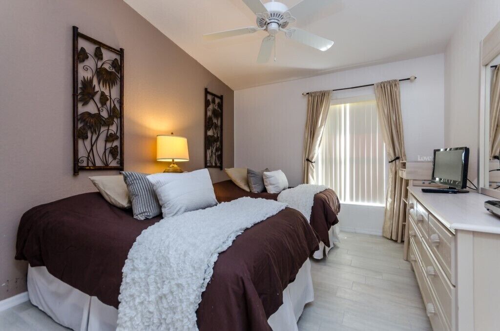 Habitación de hotel con dos camas, ventilador de techo, escritorio con televisor y ventana con cortinas.