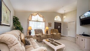 En stue med en beige sofa, to lænestole, et sofabord og et fjernsyn.