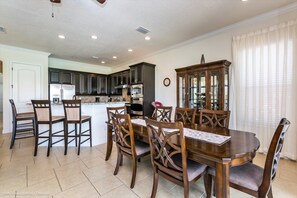 Uma sala de jantar com mesa e cadeiras de madeira, uma cozinha ao fundo e um relógio na parede.