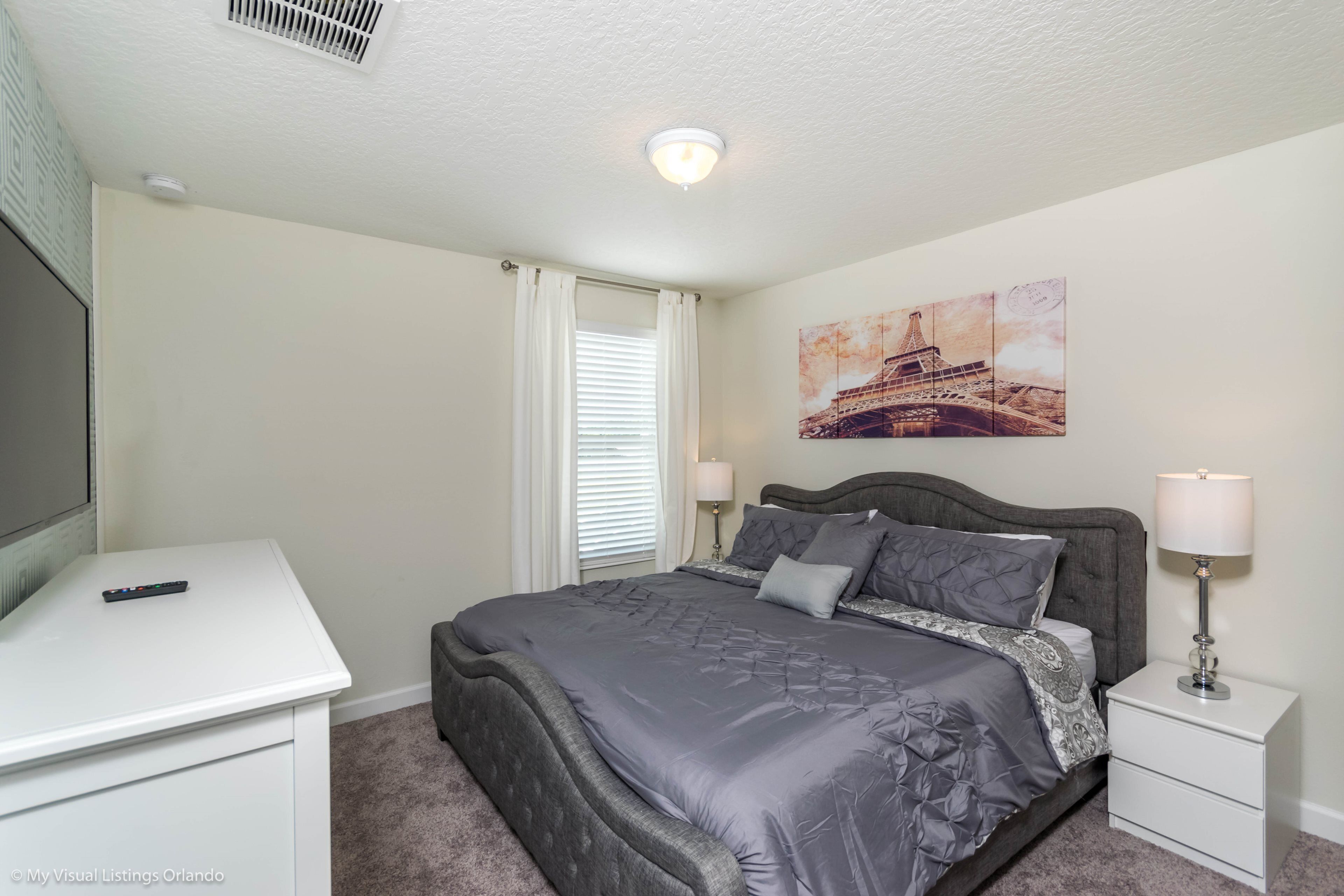 A bedroom with a bed, bedside tables, a lamp, a painting of the Eiffel Tower, and a remote control on a desk.