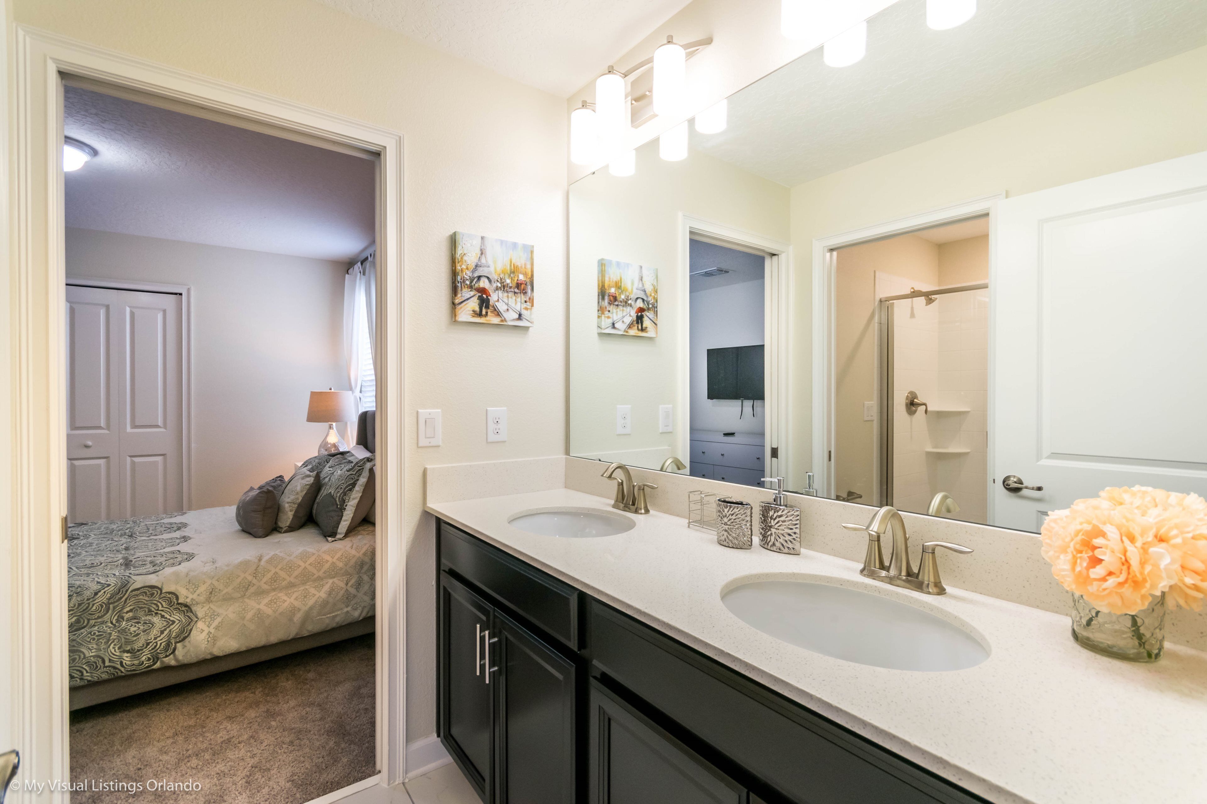A bathroom with two sinks, a large mirror, and a bed in the adjacent room.