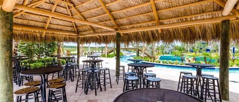 An outdoor patio area with thatched roof, multiple tables and bar stools, and a pool in the background.