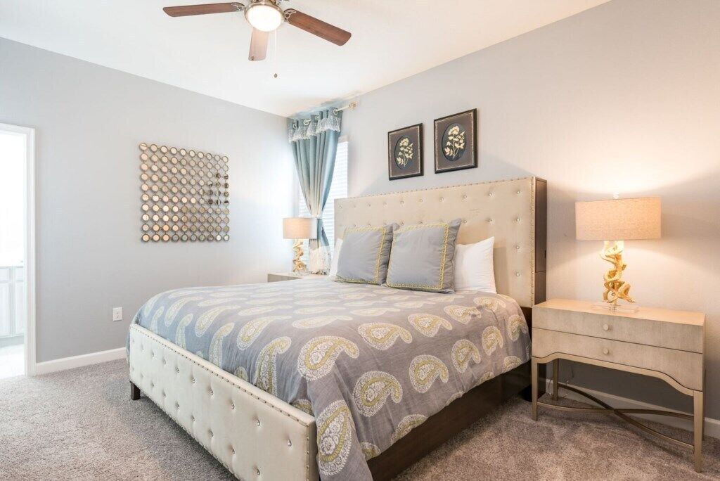 Un dormitorio con una cama de cabecera capitonada, ventilador de techo, panel decorativo en la pared y una lámpara de noche.