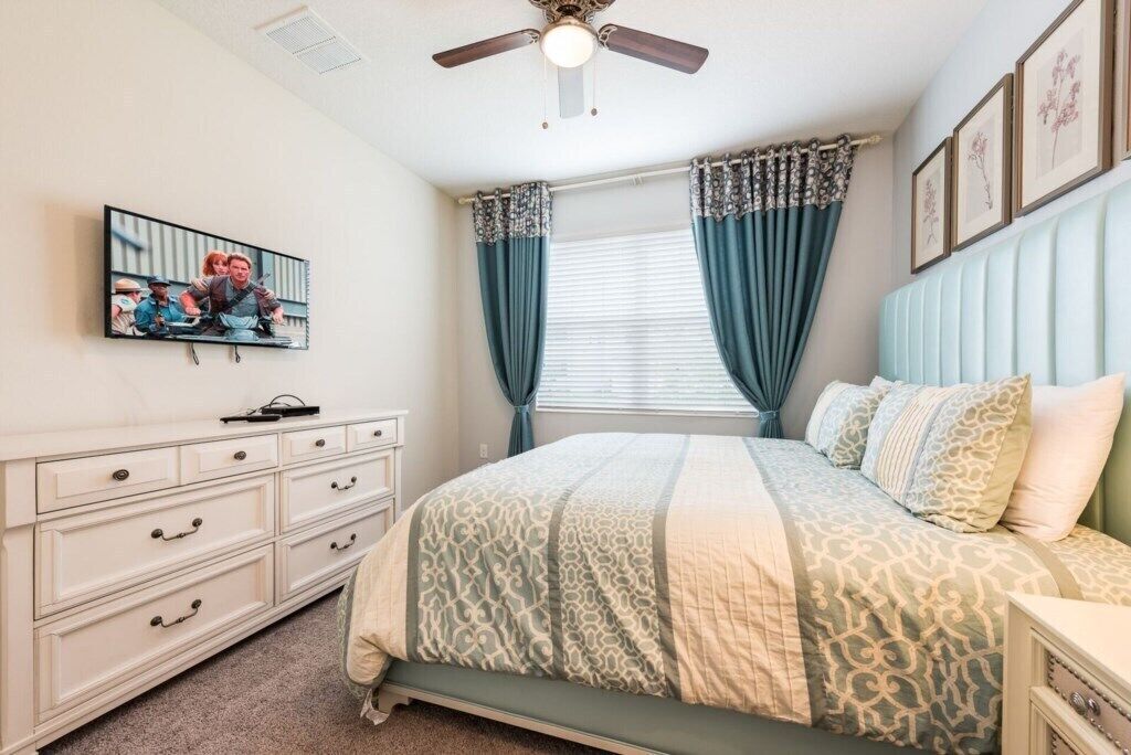 Un dormitorio con cama, cómoda, ventilador de techo, televisor y cortinas.