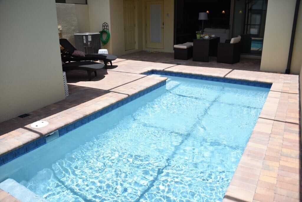 A rectangular swimming pool with a tiled edge, surrounded by a patio area with outdoor furniture and a door leading to an indoor space.