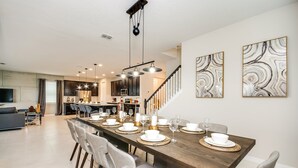 Un comedor amplio con una larga mesa de madera dispuesta para seis comensales, rodeada de sillas grises tapizadas. Se ve una cocina con armarios oscuros y una escalera en el fondo.