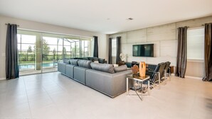 Un salón comedor amplio con un sofá seccional gris grande, un televisor de pantalla plana montado en la pared y una zona de estar con sillas y una mesa.