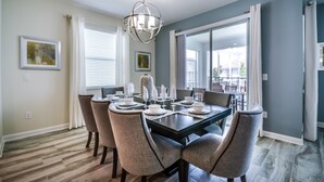 Een eetgedeelte met een tafel voor vier personen, omringd door zes stoelen, een kroonluchter en grote ramen met jaloezieën.