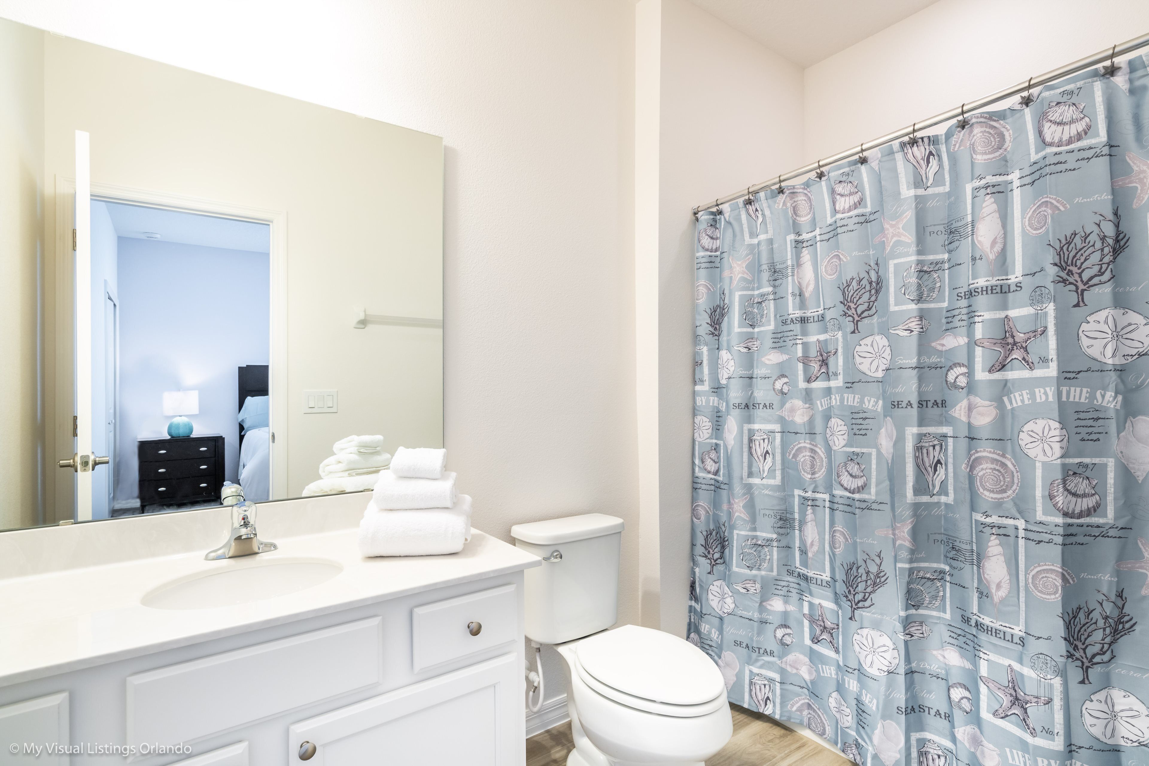 Une salle de bain avec un lavabo blanc, un miroir et des toilettes. Il y a un rideau de douche orné d’un motif de coquillages.