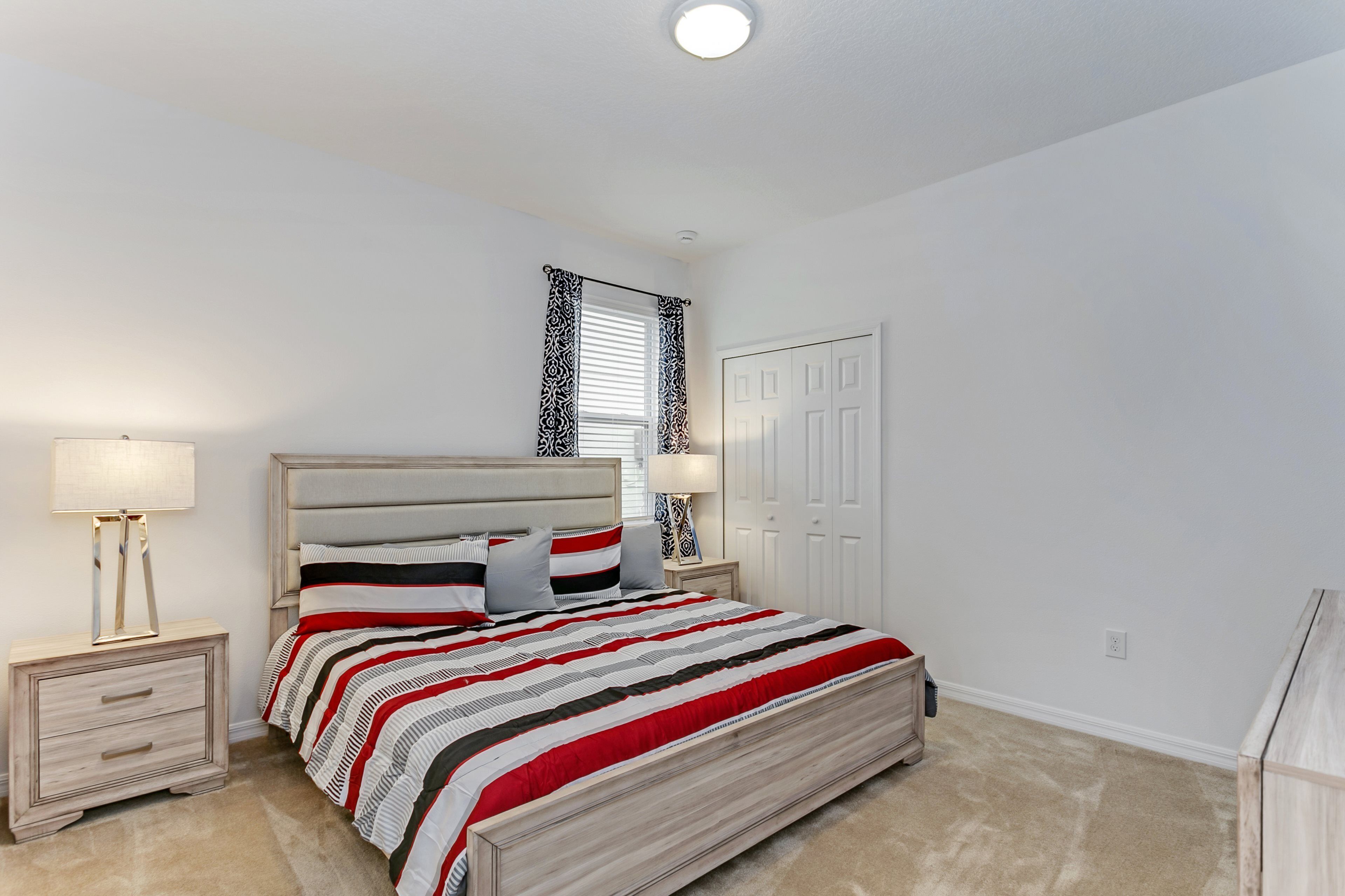 A bedroom with a bed, two bedside tables, a nightstand, a window with patterned curtains, and a door.