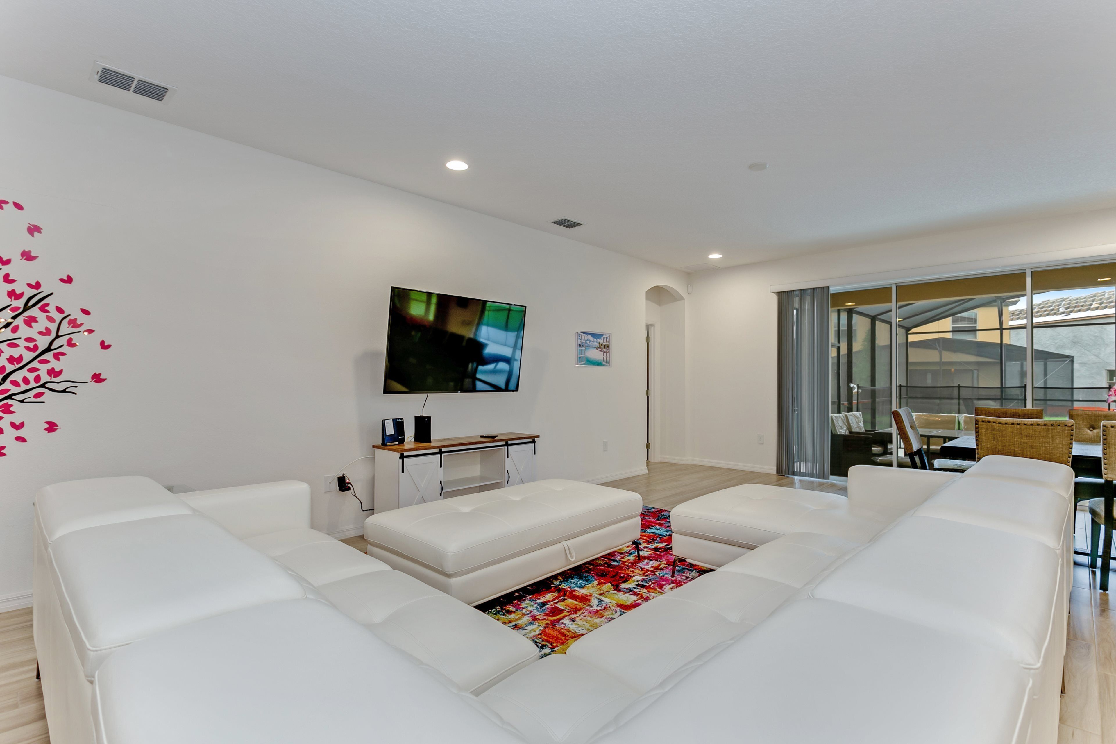 A modern living room with a large flat-screen TV, white leather sofas, a colorful rug, and a glass door leading to an outdoor area.