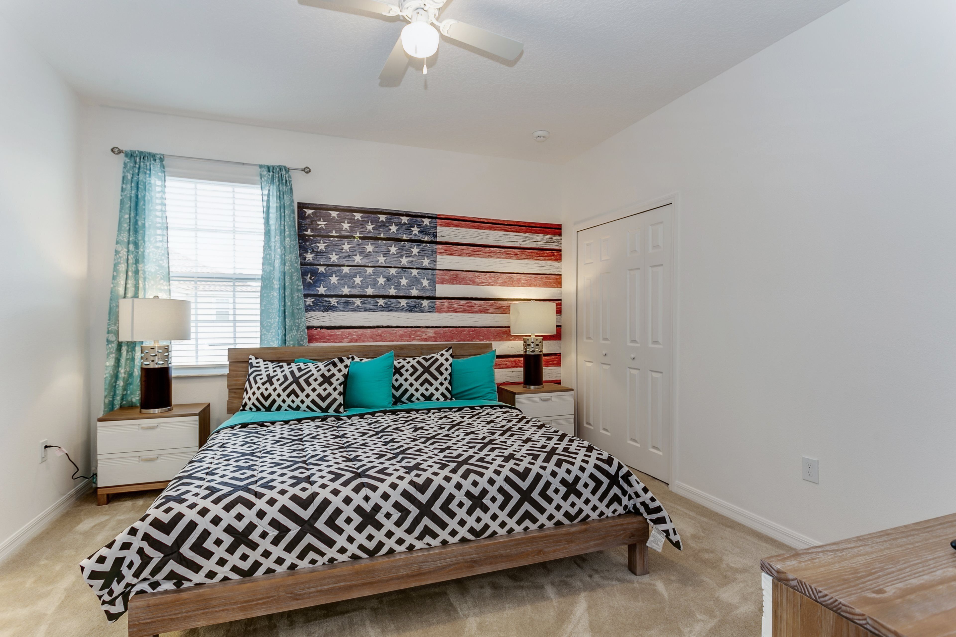 A bedroom with a bed, nightstands, a ceiling fan, a window with curtains, and a wall with a flag design.