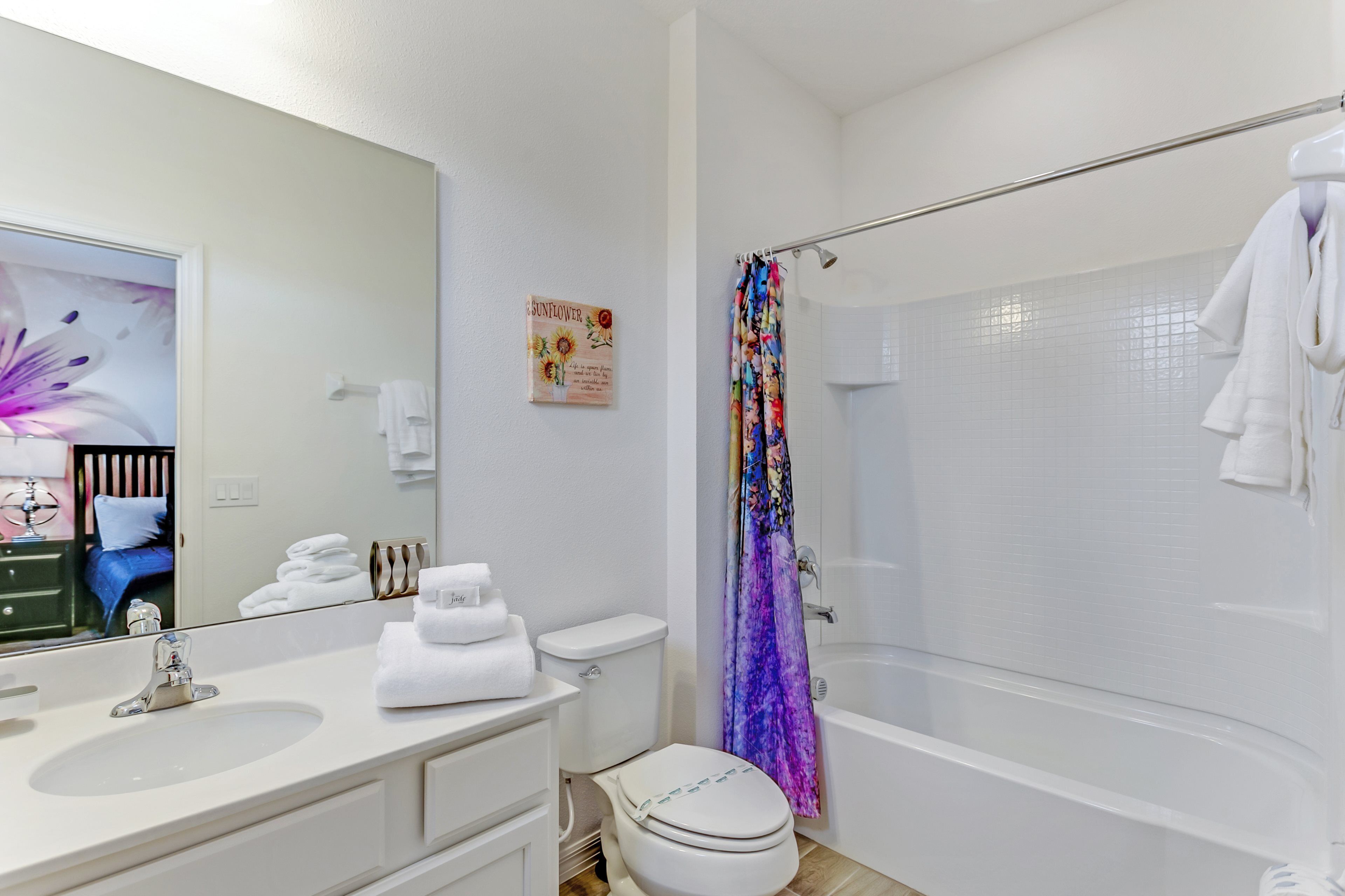 Un baño con lavamanos, inodoro y bañera blancos. Hay una cortina de ducha con estampado floral y un perchero con toallas blancas.
